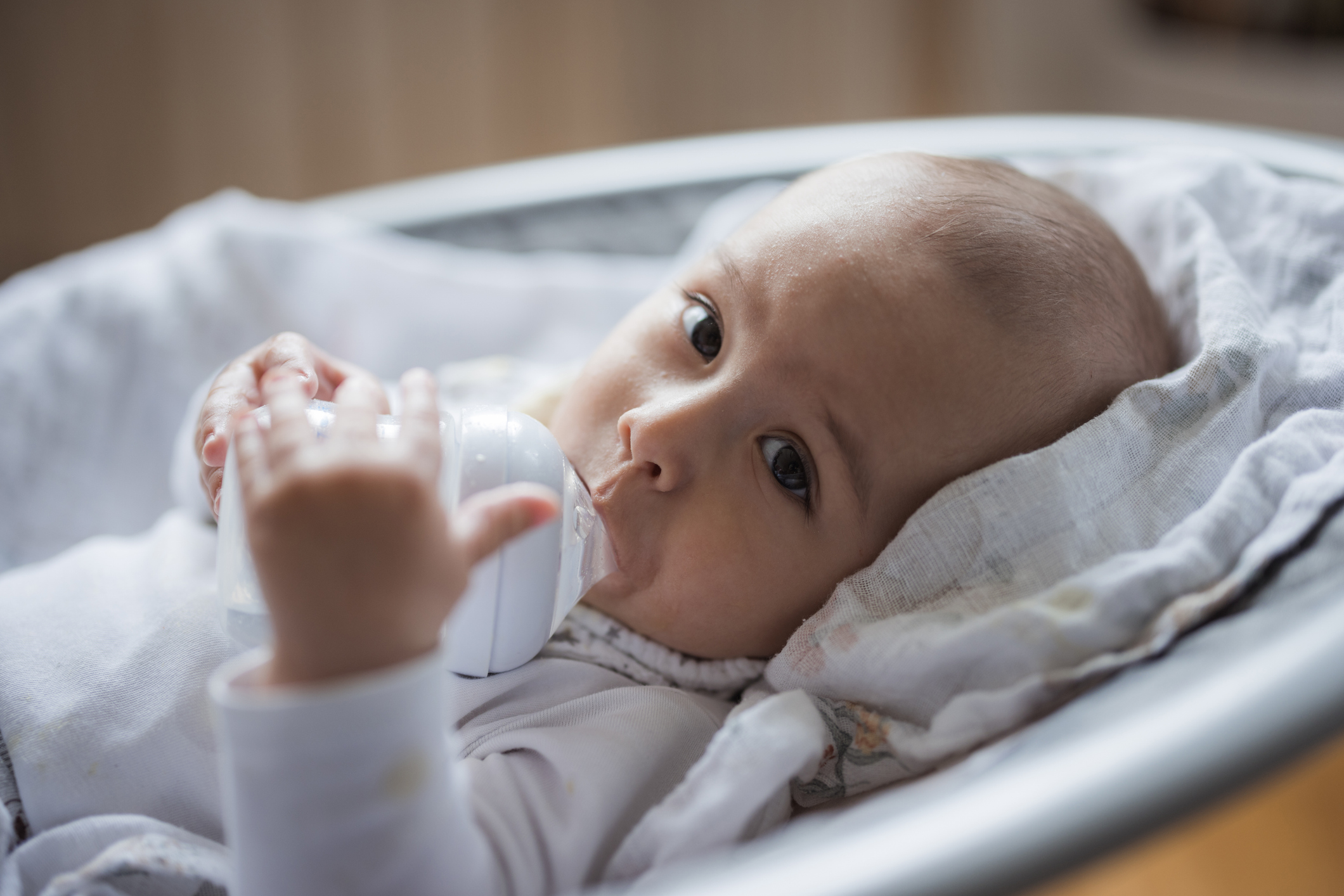 A baby drinking water from a milk bottle in a bouncer.