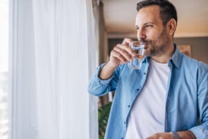 A man standing by a window, drinking a glass of water.